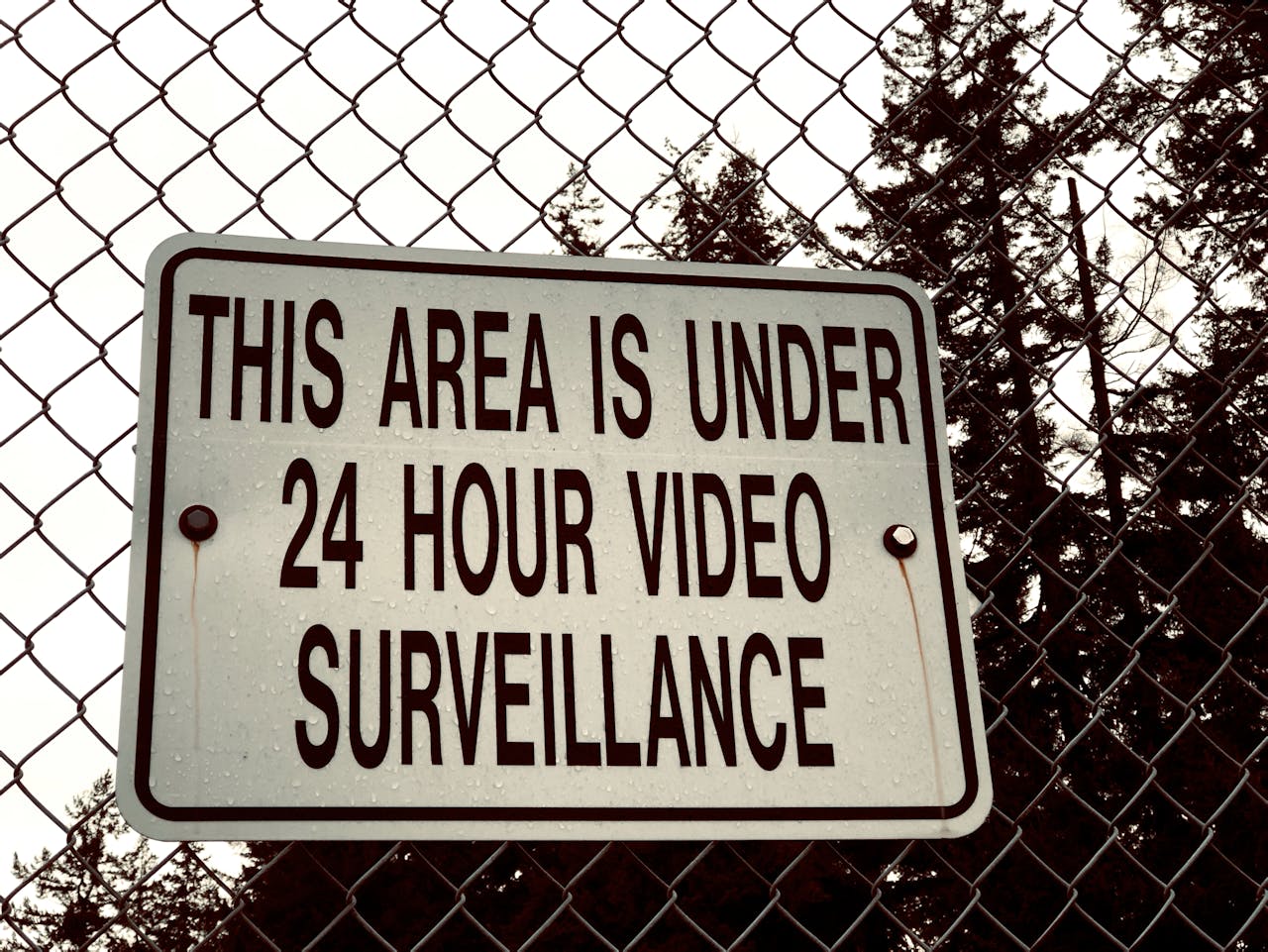 Sign on a chain-link fence stating 'This area is under 24 hour video surveillance.'